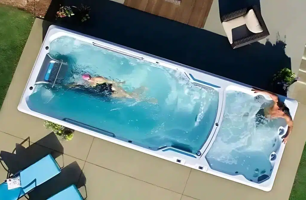 Vue aérienne d'un spa de nage rectangulaire dans un jardin ; une personne nage à gauche, tandis qu'une autre se détend dans le bain bouillonnant à droite. Des chaises de patio et une table se trouvent à proximité.
