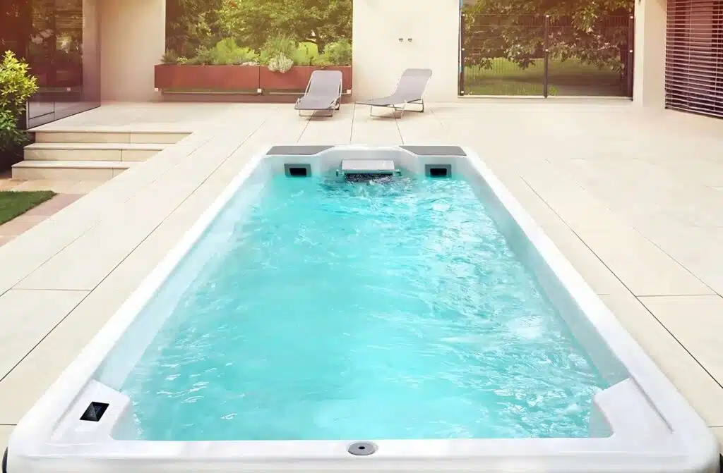 Un jardin moderne comprend une piscine rectangulaire aux eaux bleu clair, entourée de carrelage clair. Deux chaises longues sont installées au fond, près de grandes baies vitrées et d'une végétation luxuriante.