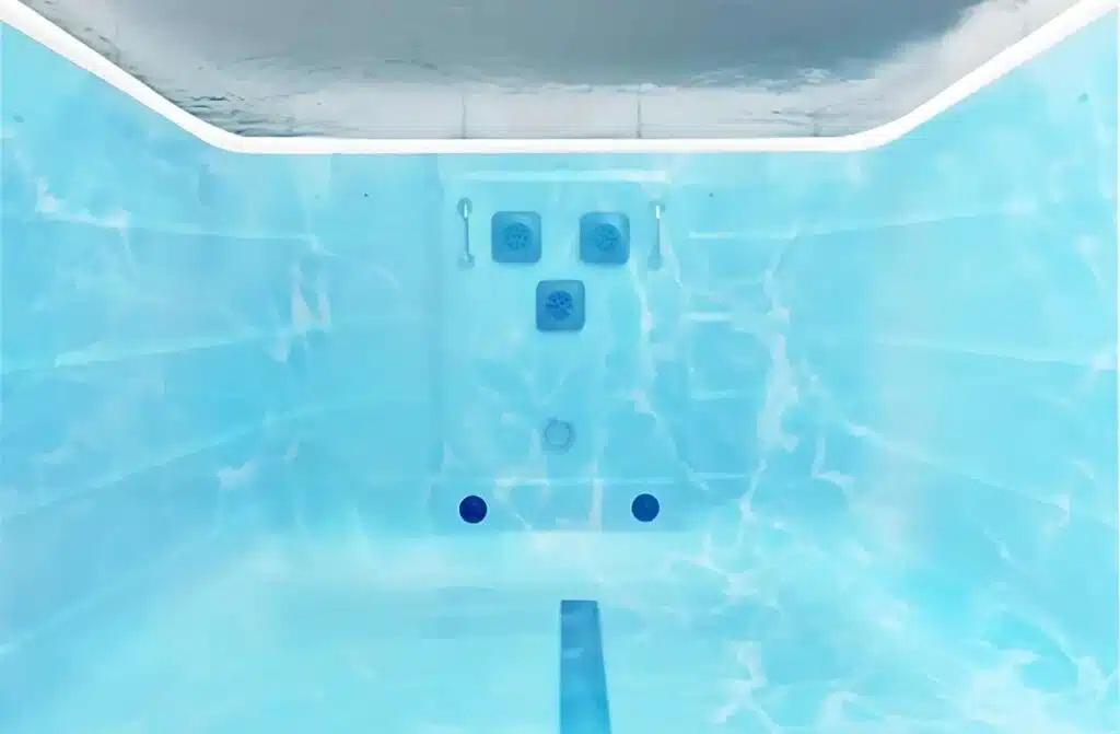 Vue sous-marine d'une piscine aux parois carrelées de bleu, avec deux poignées et des aérations visibles sur le mur du fond. La lumière venant du dessus crée des reflets ondulants sur les surfaces du bassin.
