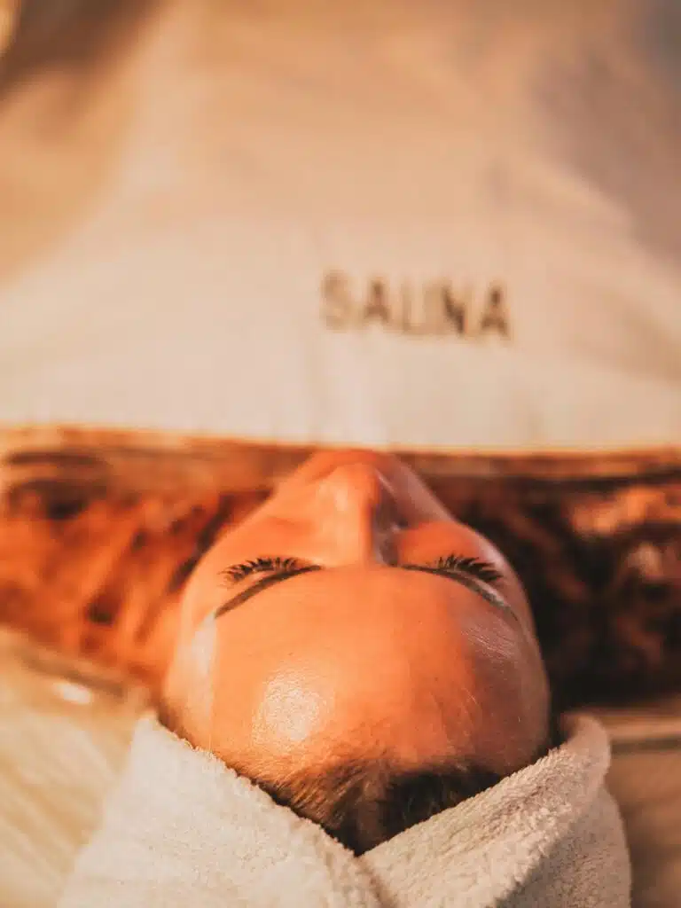 Une personne est allongée sur le dos, la tête enveloppée dans une serviette, les yeux fermés, à l'intérieur d'un espace étiqueté « Sauna ». L'éclairage est chaleureux, créant une atmosphère relaxante et sereine, illustrant parfaitement les bienfaits du sauna pour l'esprit et le corps.