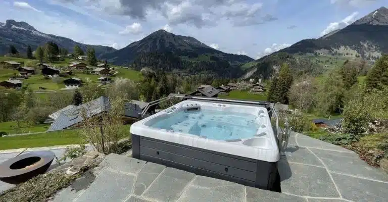 Un bain à remous se trouve sur un patio en pierre surplombant un paysage montagneux pittoresque avec des maisons dispersées et une verdure luxuriante. Le ciel est partiellement nuageux avec des taches bleues, mettant en valeur les environs sereins et pittoresques.