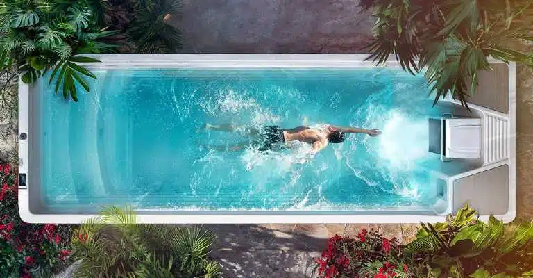 Un homme nage en nage libre dans une étroite piscine de jardin entourée de plantes vertes luxuriantes et de fleurs rouges, créant des éclaboussures dans l'eau bleue. La photo est prise d'en haut.