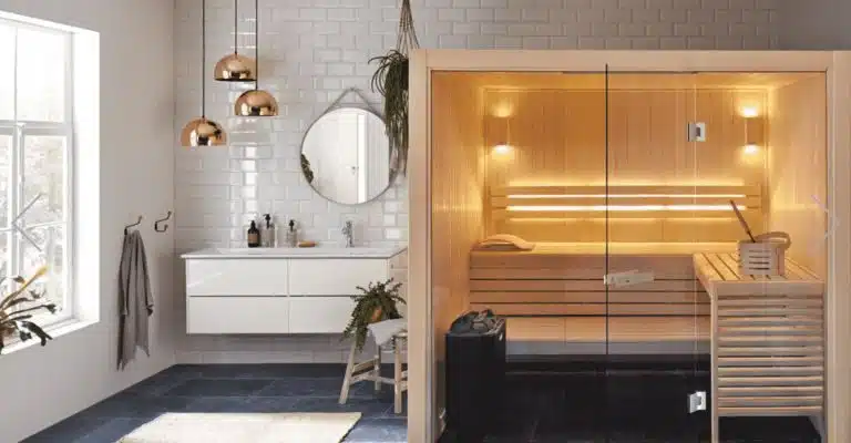 Une salle de bains moderne comprend un sauna en bois clair avec des portes vitrées sur la droite. Sur la gauche, il y a une vanité avec un miroir rond, des tiroirs blancs et des suspensions métalliques suspendues au plafond. Une fenêtre permet à la lumière naturelle d'éclairer la pièce.