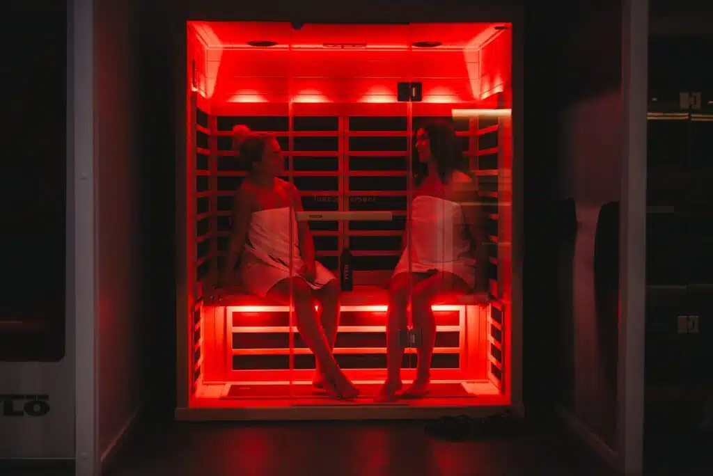 Deux personnes enveloppées dans des serviettes blanches sont assises dans un sauna infrarouge baigné de lumière rouge. Ils se font face et semblent en conversation. L’arrière-plan est faiblement éclairé avec des murs sombres, soulignant la lueur rouge vif du sauna.