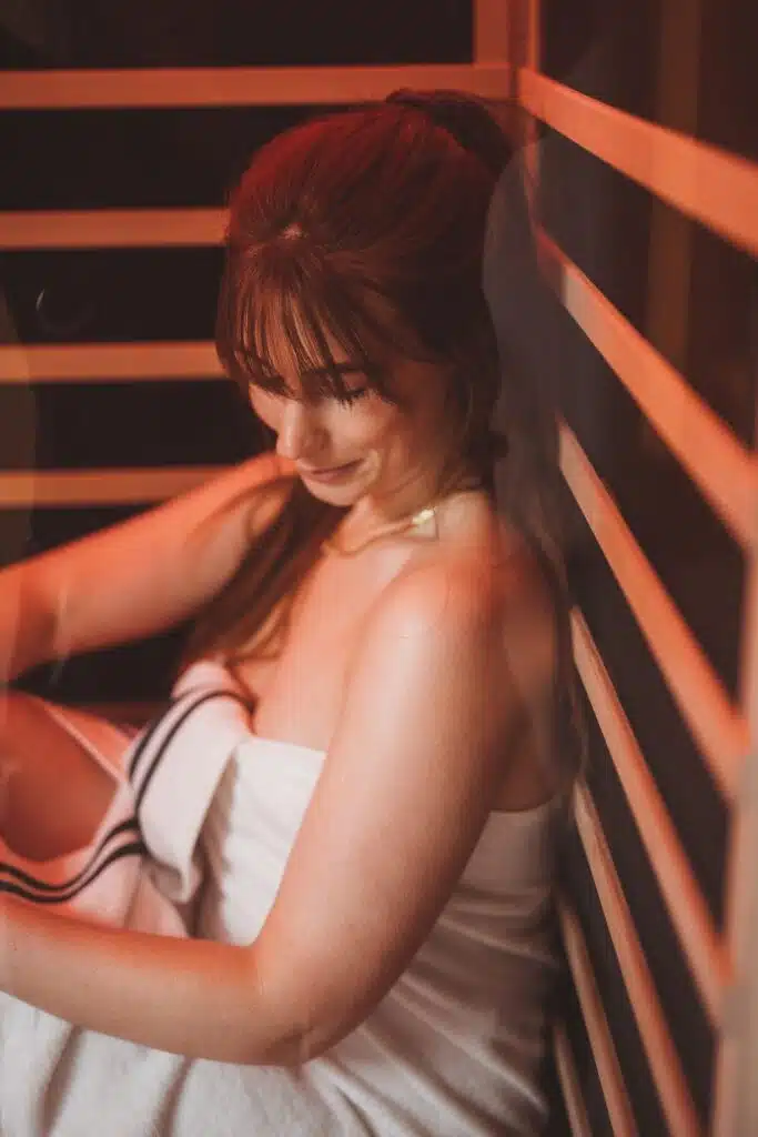 Une femme aux longs cheveux bruns est assise dans un sauna infrarouge, enveloppée dans une serviette blanche. Elle a une frange et regarde vers le bas, semblant détendue. Les lattes en bois du sauna et son éclairage rouge chaleureux créent une atmosphère chaleureuse.