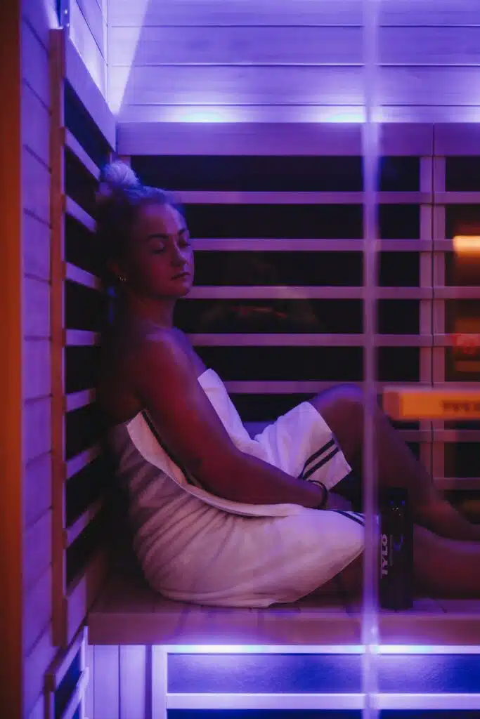 Une personne aux cheveux en chignon se détend dans un sauna infrarouge, enveloppée dans une serviette blanche. Ils sont adossés à un mur en bois baigné d’une lumière douce et violette. Une bouteille de boisson étiquetée « TOZA » est placée à côté d'eux sur le banc.