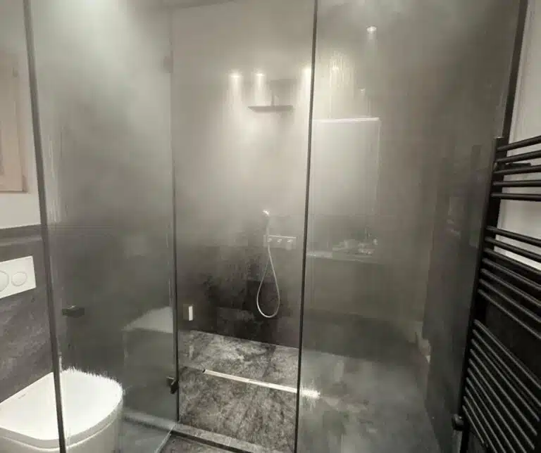 Une salle de bain moderne avec douche à l'italienne entourée de parois vitrées. La douche coule, créant de la vapeur qui embue la vitre. Il y a des toilettes blanches sur la gauche. L'intérieur présente des carreaux sombres, des luminaires élégants et un sèche-serviettes sur la droite.