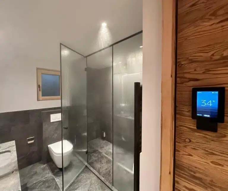 Une salle de bains moderne avec sol en marbre gris et carrelage mural. Il comprend un espace douche vitré, des toilettes blanches, une petite fenêtre et un panneau en bois avec un thermostat numérique monté sur le mur droit affichant « 34°C ». L'éclairage est minimal et chaleureux.