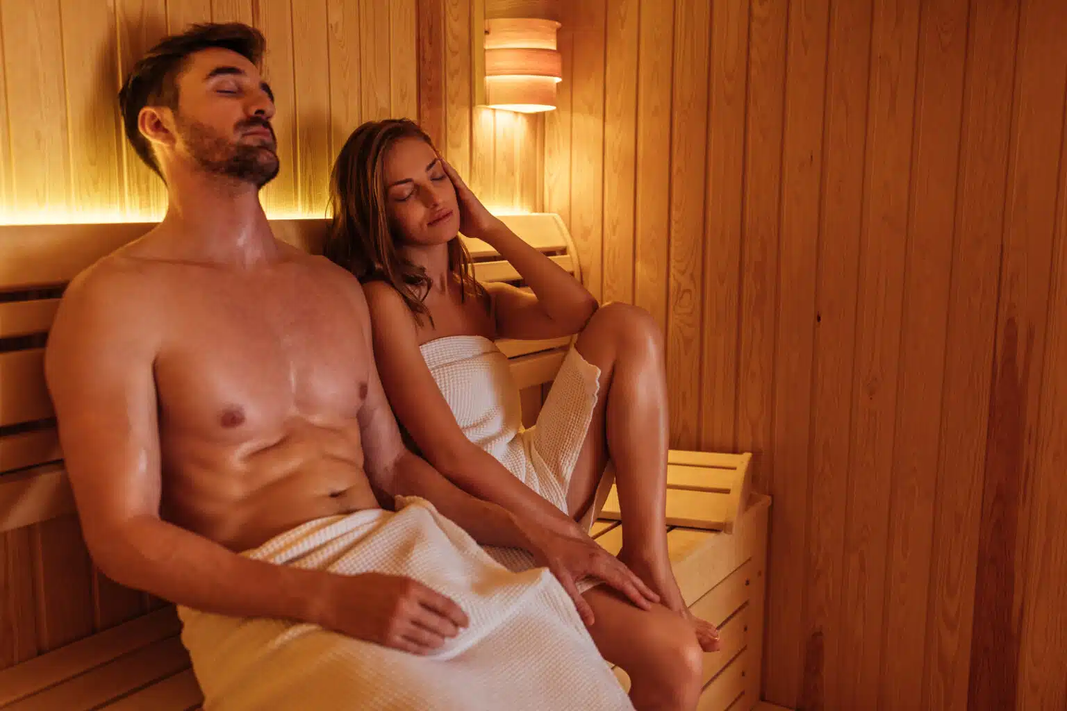 Un homme et une femme se détendent dans un sauna. Enveloppés dans des serviettes blanches, ils sont assis sur des bancs en bois, les yeux fermés, profitant de l'ambiance chaleureuse. Un éclairage doux illumine l'intérieur en bois.