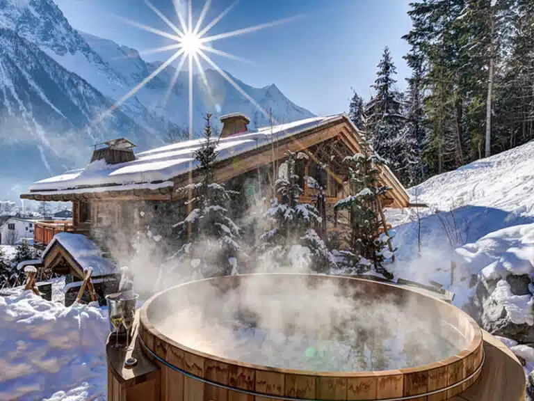 Un bain nordique en bois fumant trône dans un paysage enneigé, avec une cabane rustique en arrière-plan. La lumière du soleil brille sur les arbres et les sommets enneigés, créant une scène hivernale sereine.