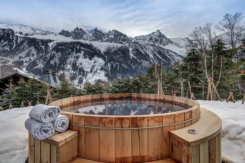 Un bain nordique traditionnel en bois avec des serviettes roulées est installé sur une terrasse enneigée surplombant une chaîne de montagnes sous un ciel nuageux. Des pins entourent la zone, créant une atmosphère sereine et isolée.