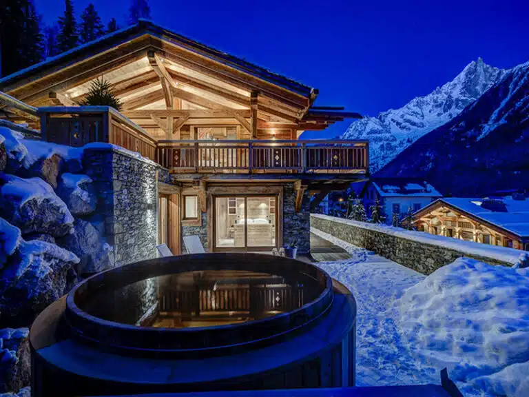 Un chalet luxueux aux lumières chaleureuses niché dans un paysage enneigé au crépuscule. La structure en bois dispose d'un balcon et d'un bain nordique accueillant. Les montagnes enneigées s'élèvent en arrière-plan, créant une scène hivernale sereine.