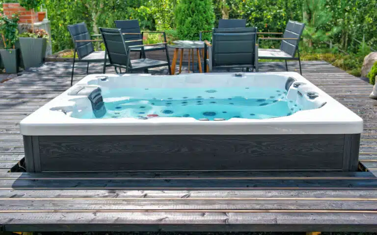 Un jacuzzi extérieur moderne est installé sur une terrasse en bois entourée de chaises de patio noires et d'une table, évoquant la tranquillité d'un sauna suisse aux normes. Le feuillage vert ajoute une toile de fond naturelle lors d'une journée ensoleillée.