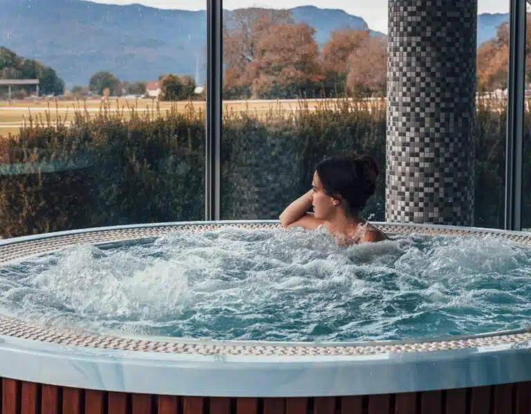 Une personne se détend dans un jacuzzi, regardant par de grandes fenêtres. La vue extérieure met en valeur les arbres et les montagnes sous un ciel nuageux rappelant les standards pittoresques que l'on retrouve dans l'expérience du sauna suisse, tandis que le décor intérieur présente des piliers carrelés et des bulles de bain tourbillonnant autour d'eux.