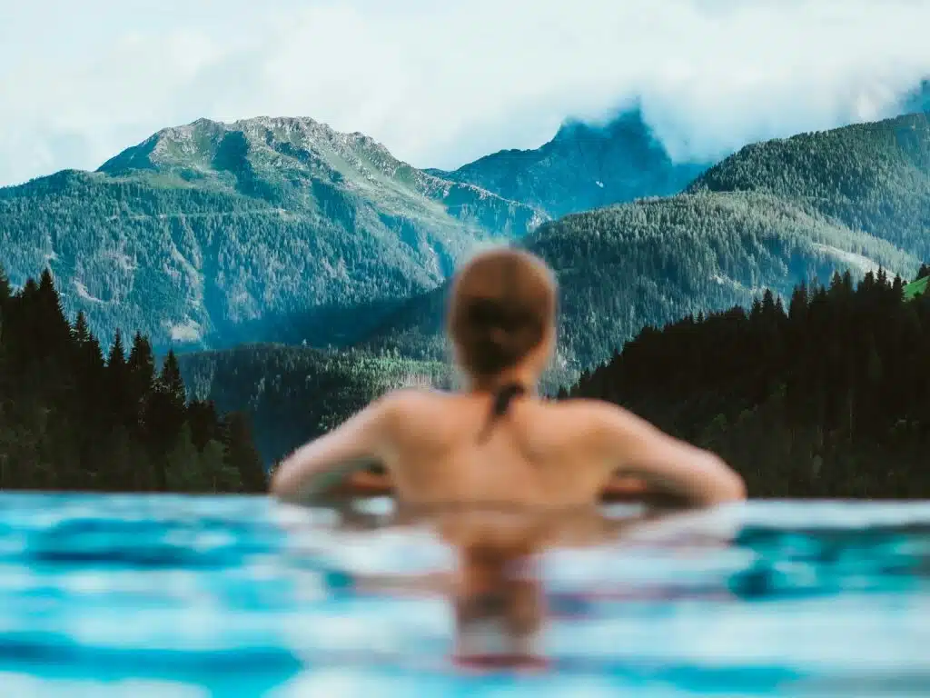 Une personne se détend dans une piscine, adossée au bord et contemplant une vue pittoresque sur les montagnes brumeuses et les forêts luxuriantes, rappelant l'ambiance tranquille que l'on trouve en Suisse. Le contraste serein entre l'eau claire et le paysage verdoyant offre une paix proche des expériences de sauna classiques.