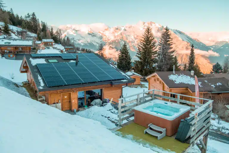 Un paysage de montagne enneigé avec des chalets en bois, dont un avec des panneaux solaires sur son toit. Au premier plan, un jacuzzi fumant offre une expérience luxueuse semblable à celle d'un sauna, entouré d'une terrasse en bois. Les sommets enneigés s'élèvent majestueusement à l'arrière-plan sous un ciel suisse clair.