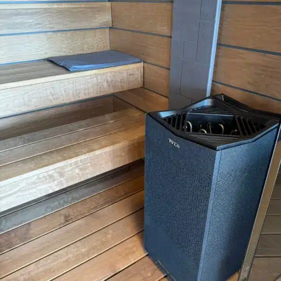 Le sauna cabine Tylö Reflection (occasion) présente un intérieur luxueux en bois avec des sièges à gradins et un élégant poêle Tylö dans un coin. Un coussin gris douillet sur la banquette supérieure renforce l'atmosphère élégante et relaxante, s'harmonisant parfaitement avec le boîtier métallique foncé du poêle.