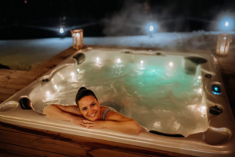 Une personne aux cheveux noirs sourit et s'appuie sur le bord d'un sauna bouillonnant la nuit. La scène est chaleureusement éclairée par des lanternes sur une terrasse en bois, avec de la brume s'élevant de l'eau, créant une atmosphère chaleureuse.