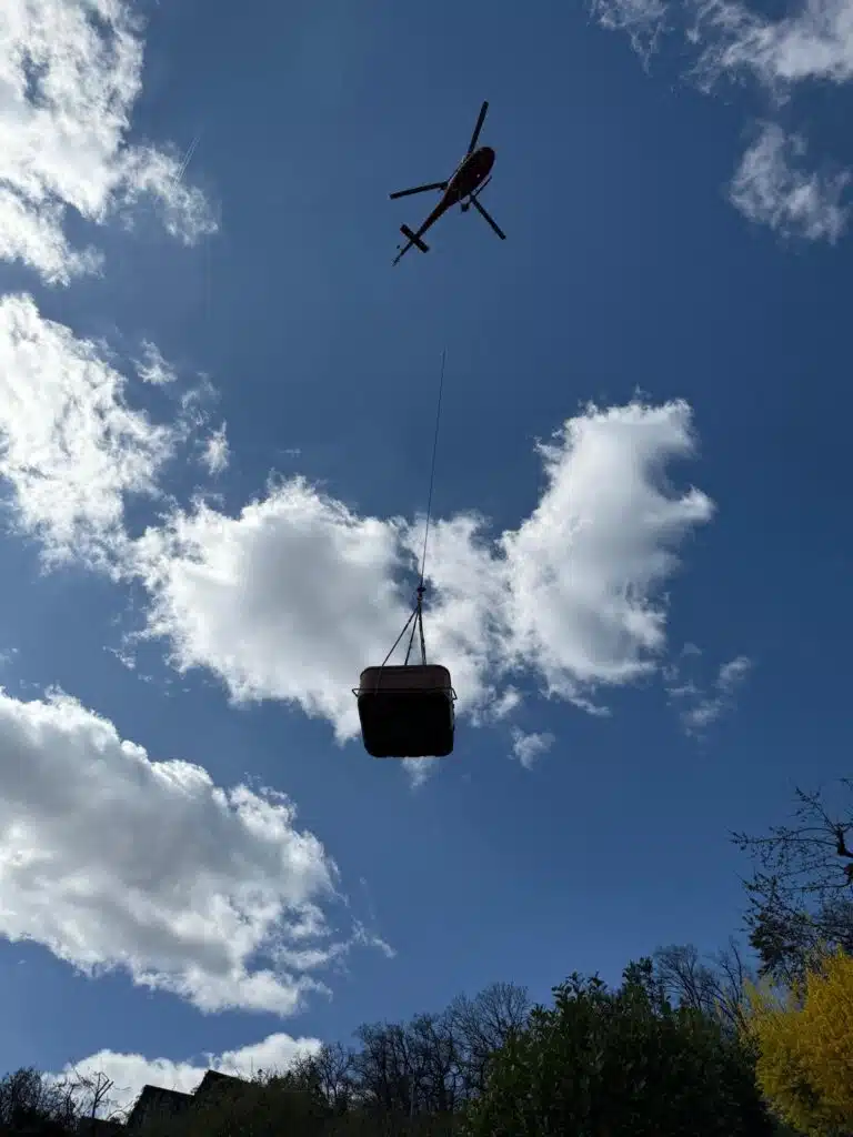 Un hélicoptère survole le paysage, se découpant sur un ciel bleu partiellement nuageux, transportant un grand conteneur suspendu à un câble. Des arbres et des arbustes sont visibles au bas de l'image.