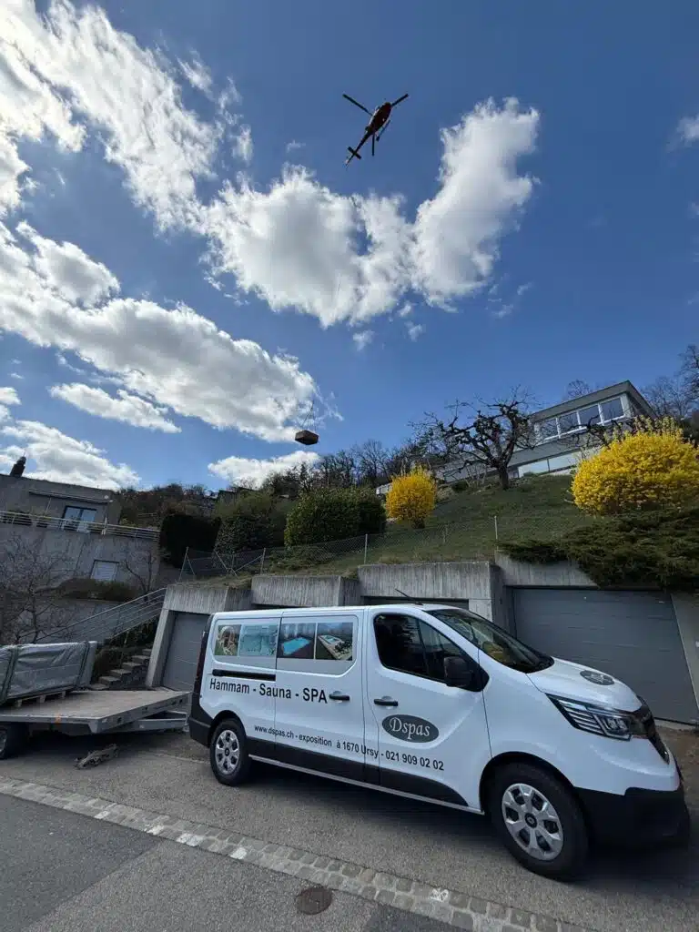 Une camionnette portant l'inscription « Hammam - Sauna - SPA » est garée dans une rue résidentielle. Au-dessus, un hélicoptère soulève un gros objet. La scène se déroule sous un ciel nuageux, avec une colline et des maisons en arrière-plan.