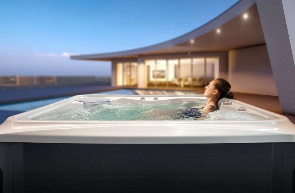 Une femme se détend seule dans un jacuzzi sur une terrasse moderne au coucher du soleil, avec une maison contemporaine et une piscine en arrière-plan, profitant de la chaleur d'une pompe à chaleur efficace.