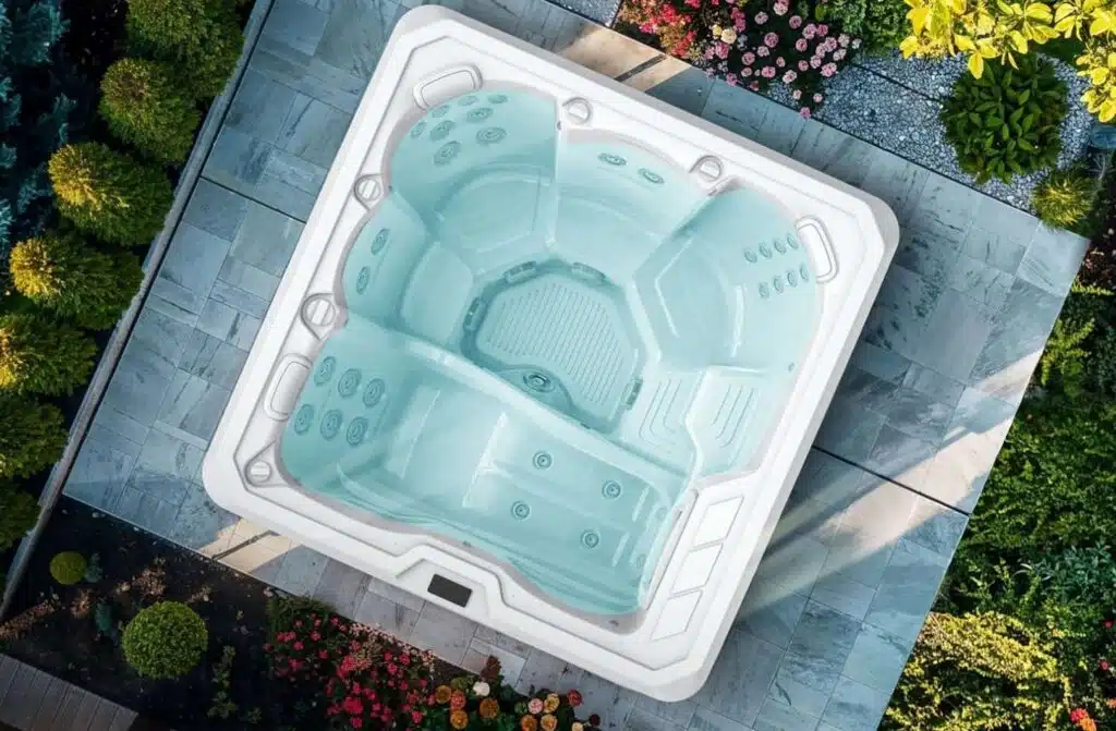 Vue de dessus d'un jacuzzi blanc vide avec des sièges moulés, installé sur un patio carrelé et entouré d'arbustes verts, d'arbres et de plantes à fleurs - une retraite de jardin sereine parfaite pour se détendre dans des spas avec une pompe à chaleur.