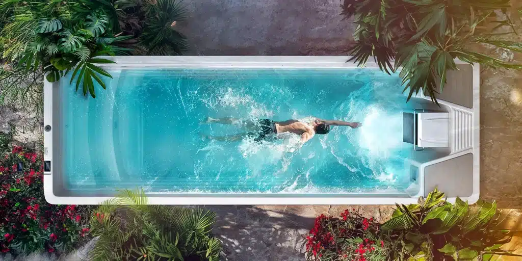 Une personne nage en nage libre dans le Spa de nage D600N rectangulaire et bleu clair, entouré de plantes vertes luxuriantes et de fleurs rouges, vu d'en haut.